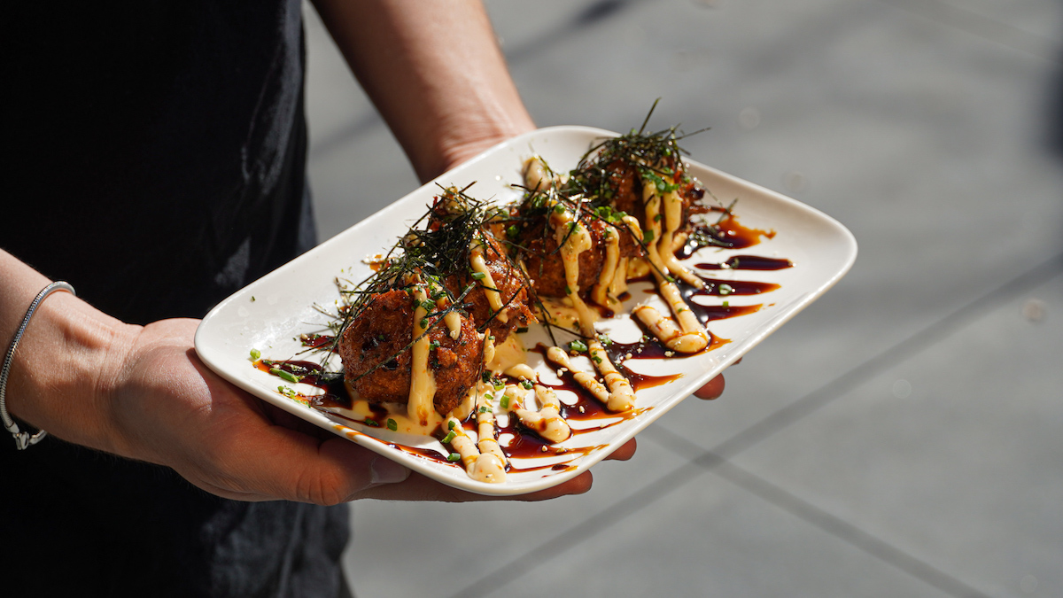 a server holds a white plate with four fried meatballs garnished with green sprouts and yellow and black sauces