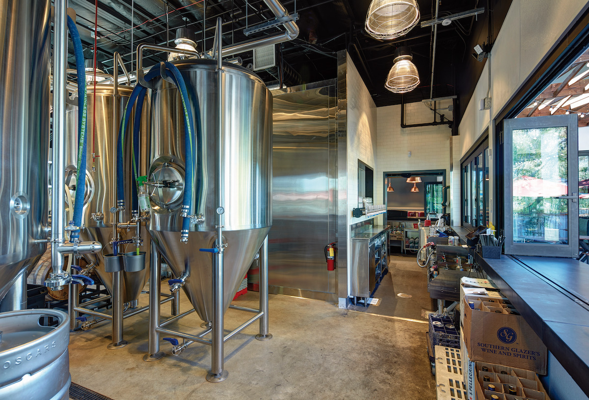 the brewing facility at Oscar's Brewing Co. featuring several metal brewing containers and metal barrels with a window to the outdoor patio to the right