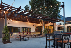 the outdoor patio of Oscar's Brewing Co. with several wooden tables and black metal chairs, a low bar top with red leather chairs facing the outdoor bar, and string lights strung above the tables