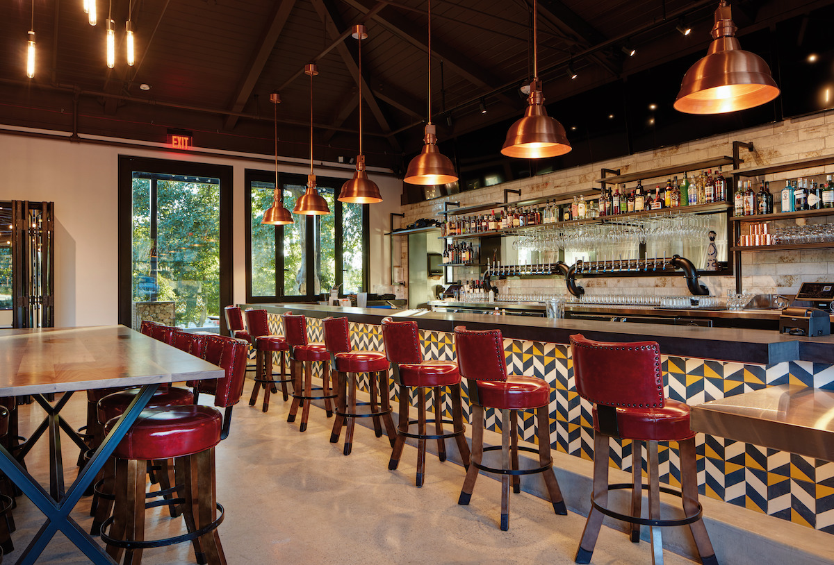 one wooden table and a bar top with red leather hightop chairs sitting in front of the bar with metal lights above the bar and beer taps and alcohol bottles behind the bar