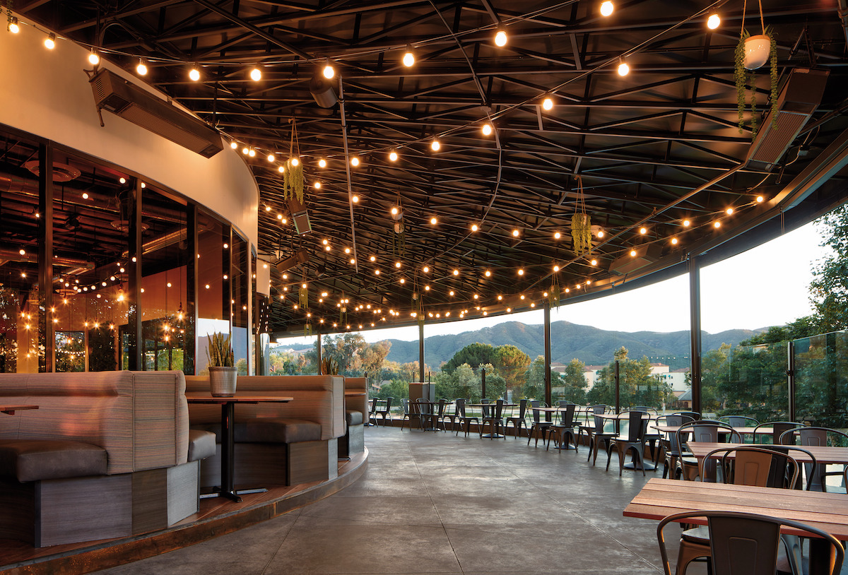 the outdoor patio at Oscar's Brewing Co. with large grey booths along the left and shorter wooden tables with metal chairs to the right overlooking the pond and mountains