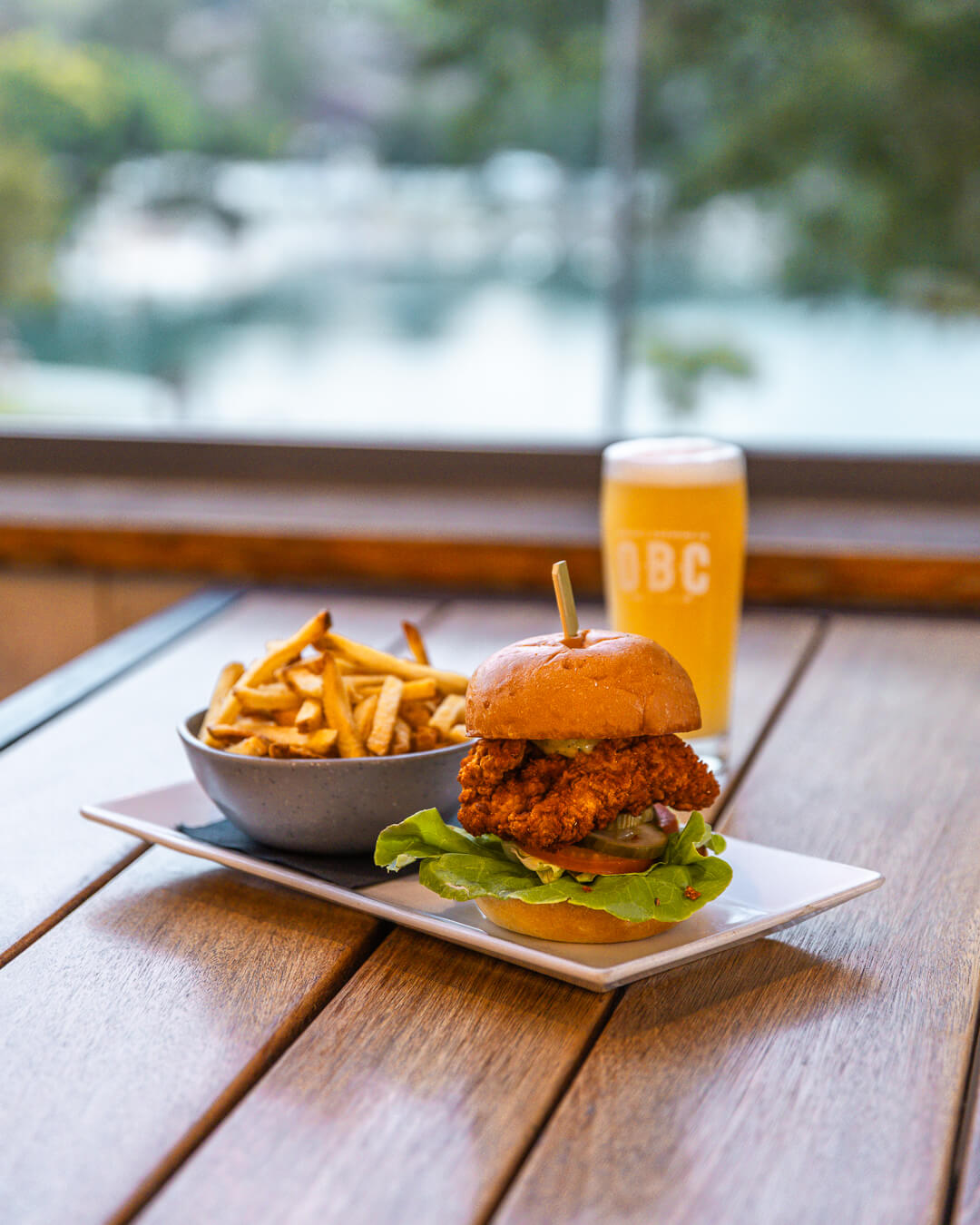 A gourmet burger with fries and a pint of beer served on a wooden patio table overlooking the water.