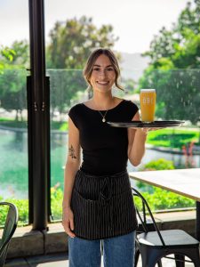 A smiling restaurant server carries a freshly poured pint of beer on a tray in an outdoor dining area.