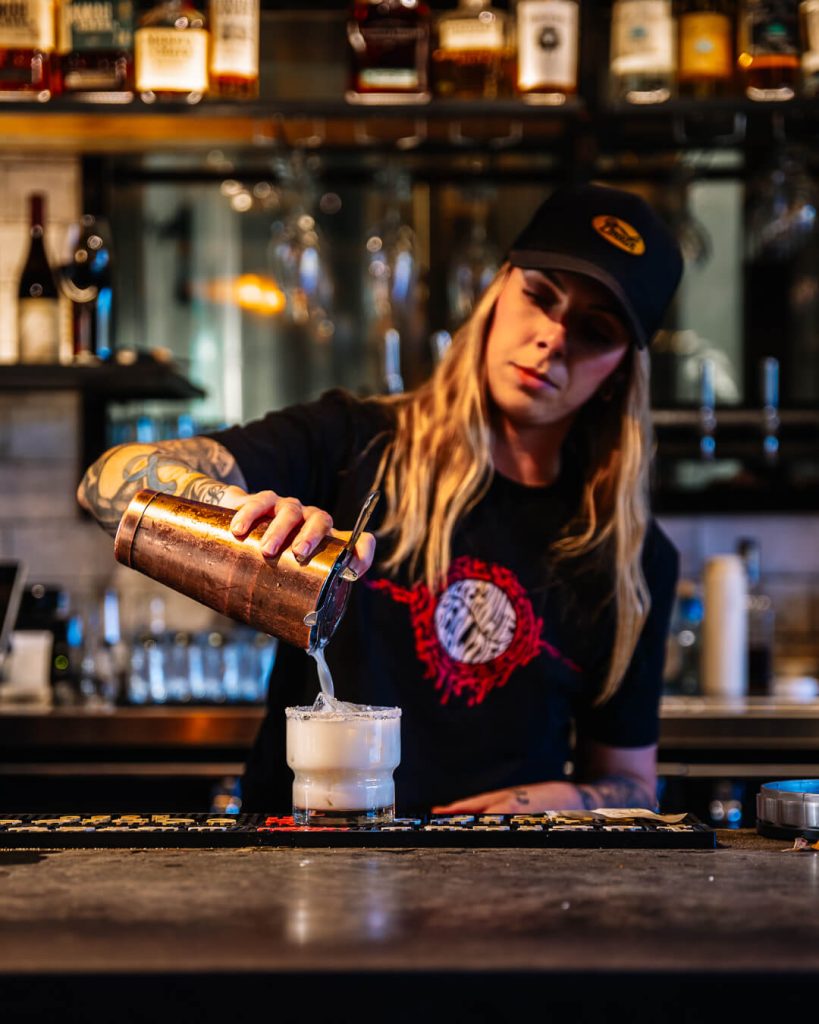A bartender prepares a handcrafted cocktail behind a fully stocked restaurant bar.