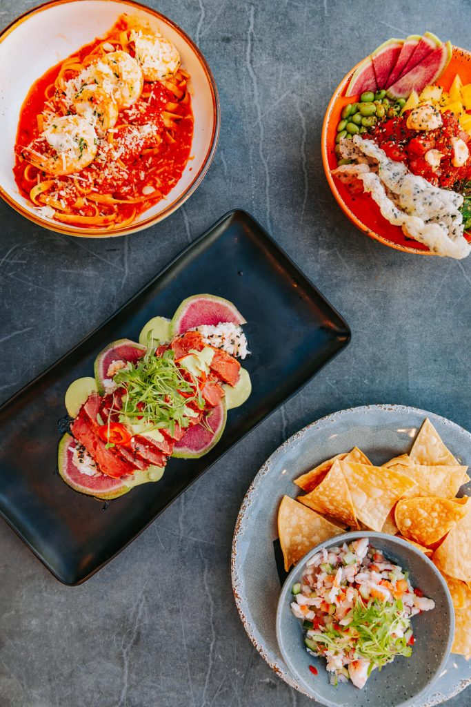 An overhead view of assorted appetizers, pizza, tacos, and fresh dishes served on a restaurant table.