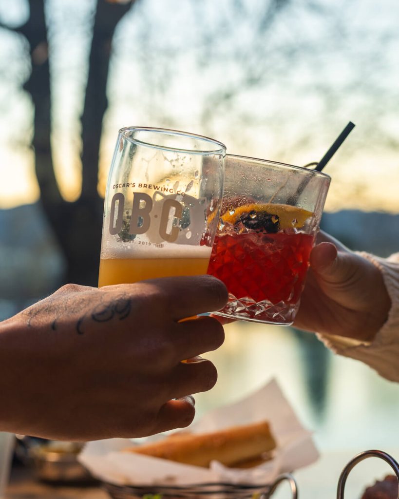 Two people clink glasses of beer and a mixed drink during an outdoor gathering at sunset.