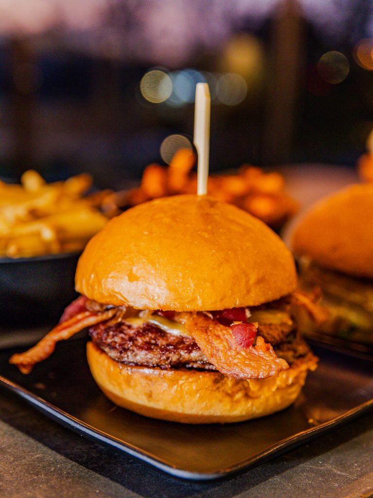 A gourmet cheeseburger topped with bacon, served on a toasted bun with fries in the background.